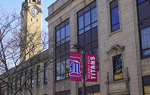 A photo of the Engineering Building near the clock tower with red banners that feature the 쿪ֱֳ logo and World Needs Titans on it.